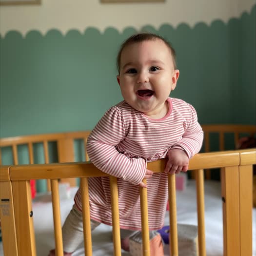 Baby Edith at 8 months old with a big smile on her face standing in her cot